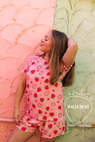 A woman wearing a pink strawberry-print pj set, posing against a split pink and green textured wall. A logo for 'MALIA IN XS' is visible in the bottom right corner.