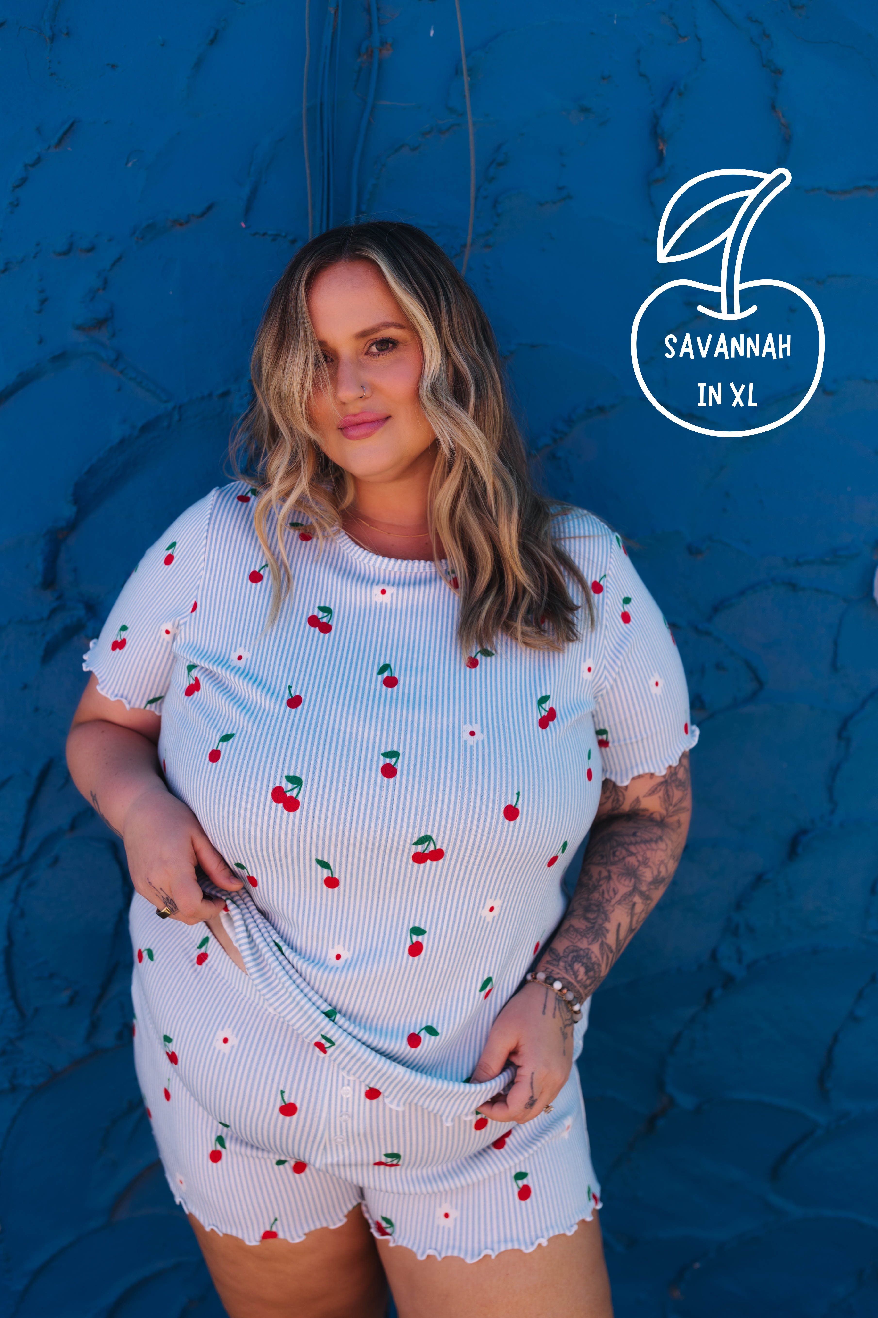A woman wearing a light blue ribbed short-sleeve top and matching shorts with a red cherry print, posing against a textured blue wall. A white circular logo in the top right reads 'SAVANNAH IN XL'.