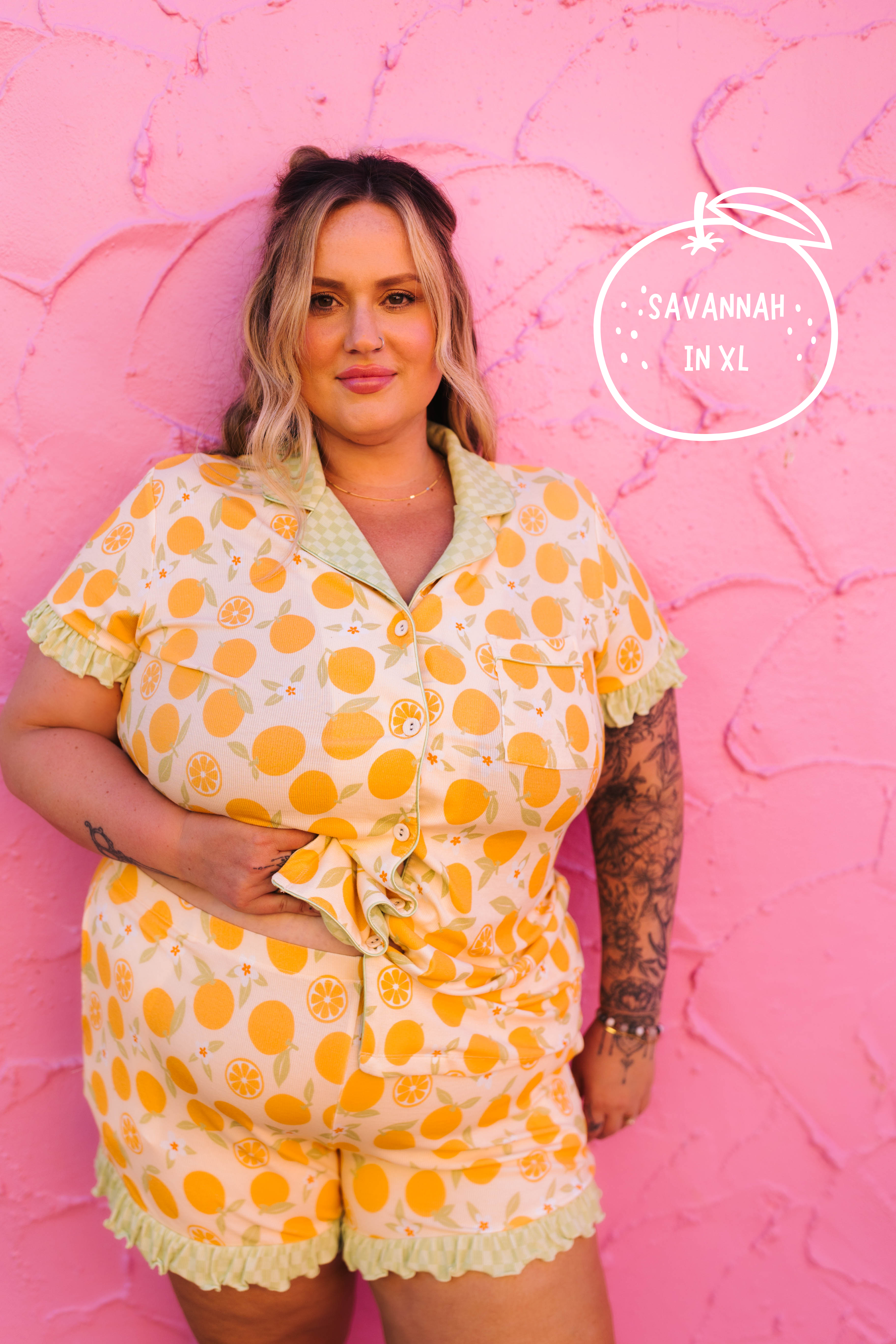 A woman modeling an orange print short-sleeve pajama set with a ruffled collar, button front, and matching ruffled shorts, standing against a textured pink wall. A logo watermark reading 'SAVANNAH IN XL' is visible on the upper right.