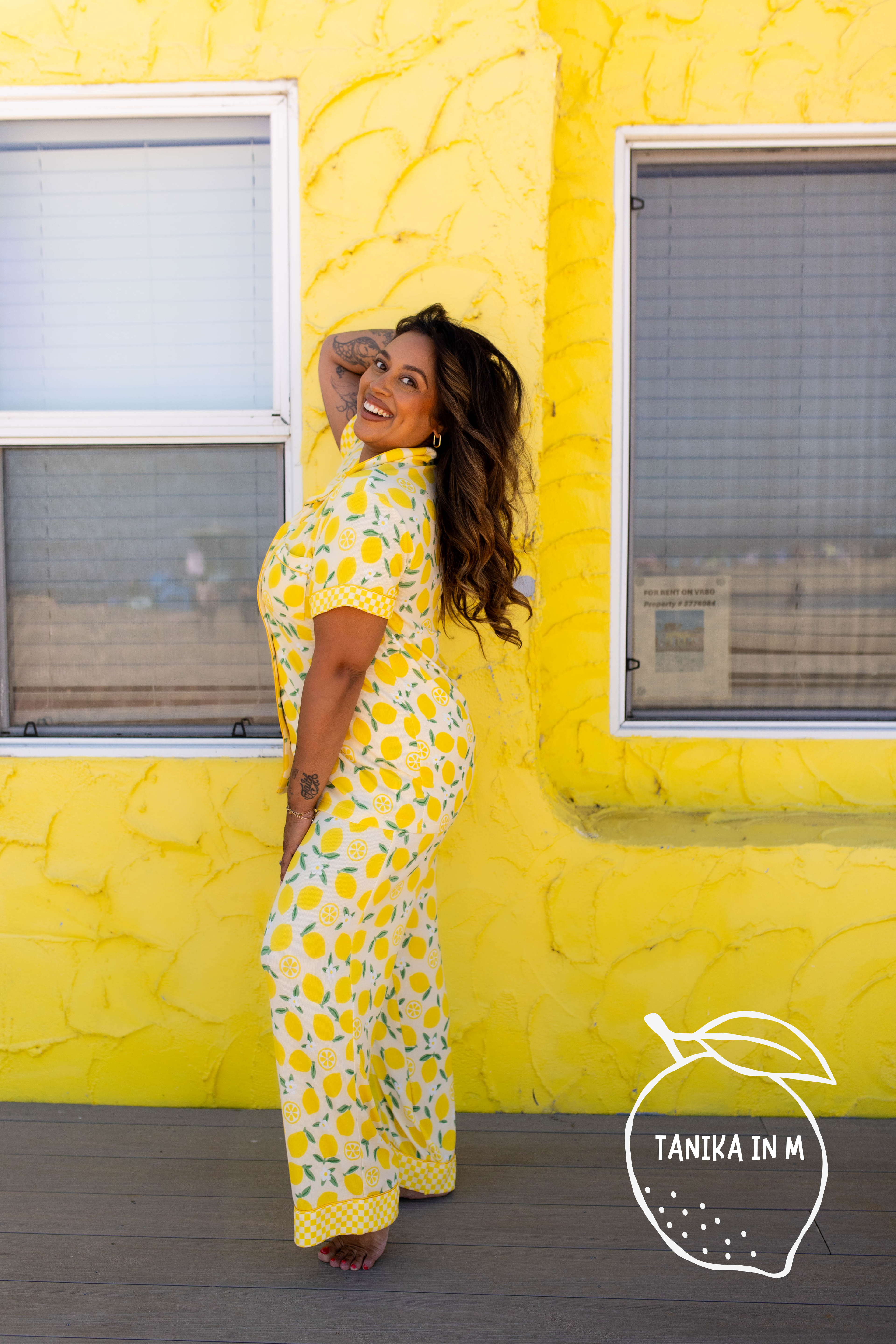 A woman posing in a yellow and white lemon-print pj in front of a bright yellow textured wall.
