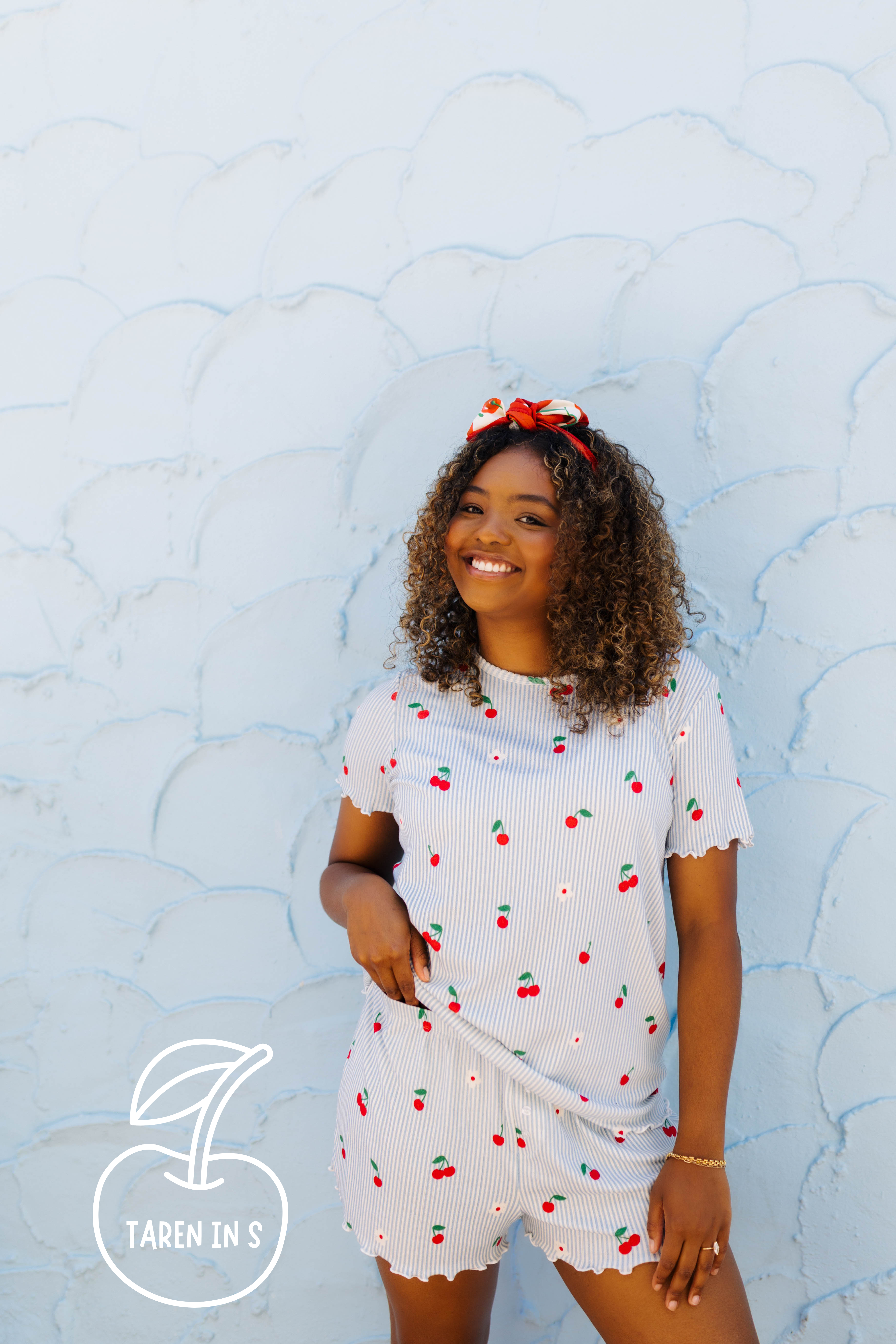 A woman wearing a white ribbed short-sleeve pj set with a cherry print, standing against a light blue textured wall. She has a red bow in her hair, and a logo for 'TAREN IN S' is visible in the bottom-left corner.