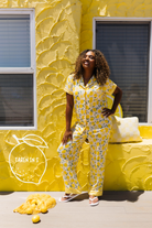 A woman modeling a yellow lemon-print pajama set stands in front of a textured yellow wall with windows. A pile of lemons and a small sign reading 'TAREN IN S' are on the ground nearby.