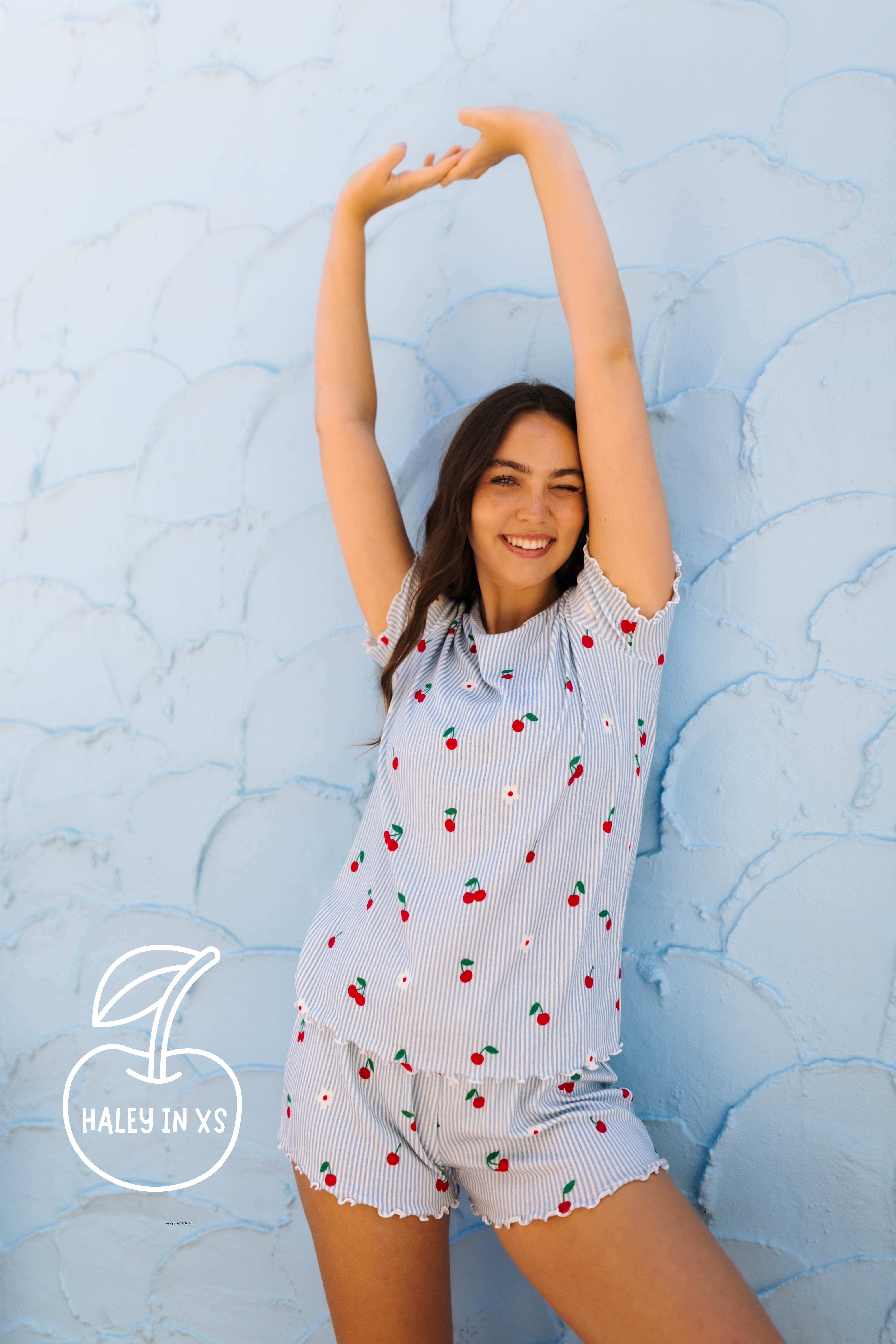 A woman wearing a light blue and white striped short-sleeve pajama set with a cherry print, posing against a textured light blue wall. A logo overlay in the bottom left reads 'HALEY IN XS'.