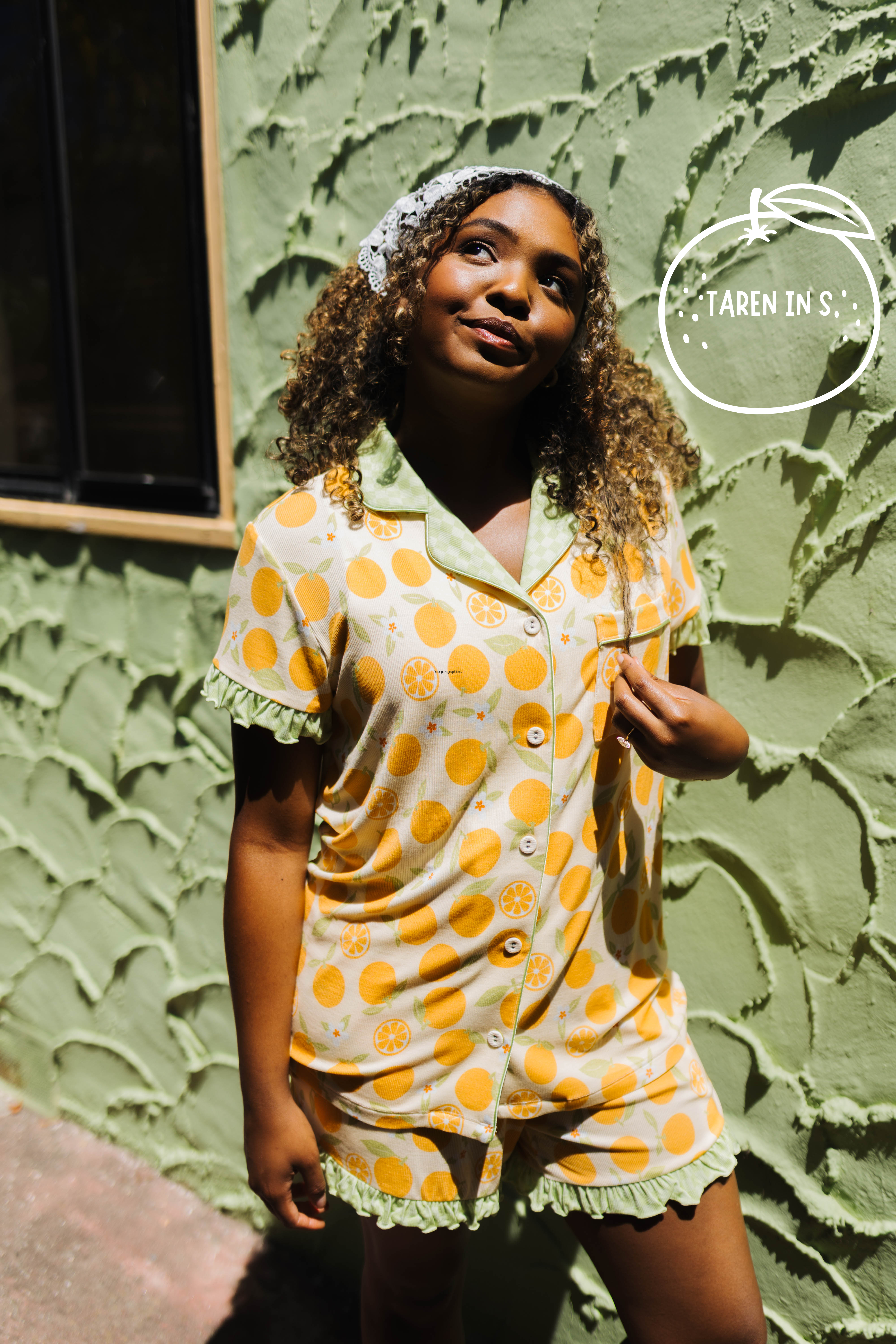 A woman wearing a two-piece pajama set with a orange citrus pattern on a cream background, standing against a textured green stucco wall. She is wearing a matching patterned headband.